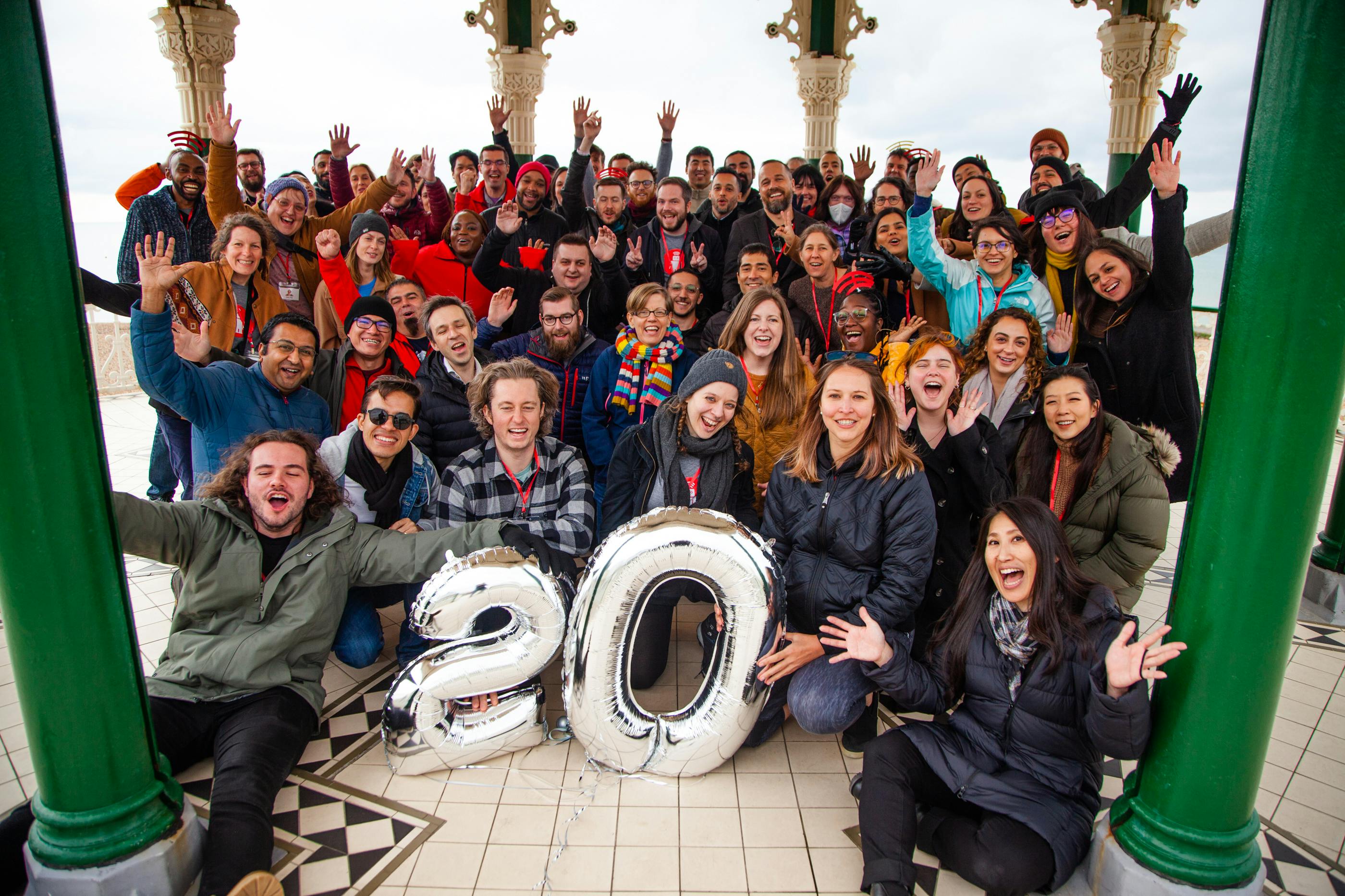 A large group of people all lined up and smiling with their hands in the air. There are two silver balloons in the front that are shaped like the number 20.