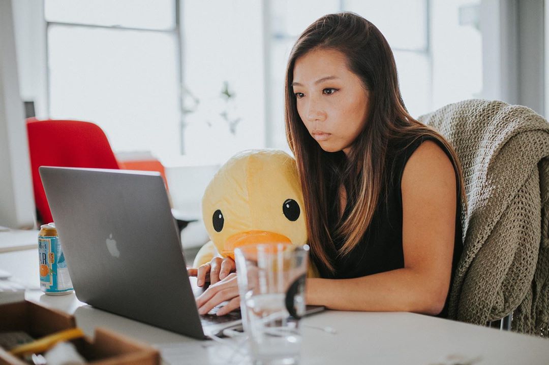 A woman holding an oversized plush duck sitting at a desk in an office, looking at a laptop.