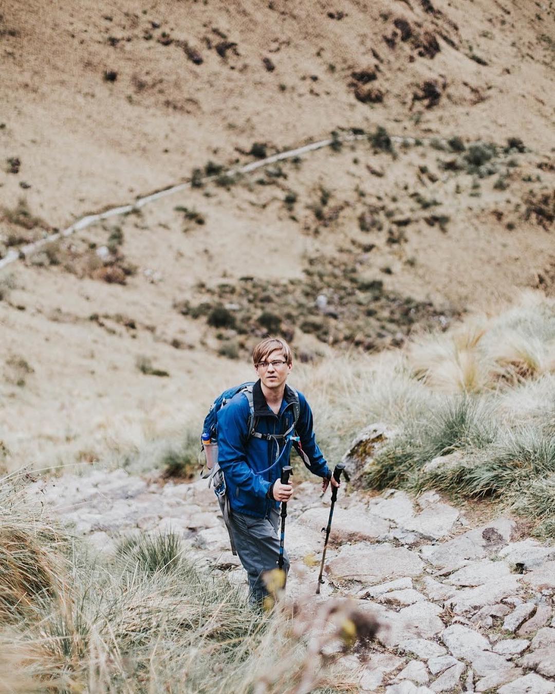 A man in a blue jacket and hiking gear looking to the side on a mountain path.